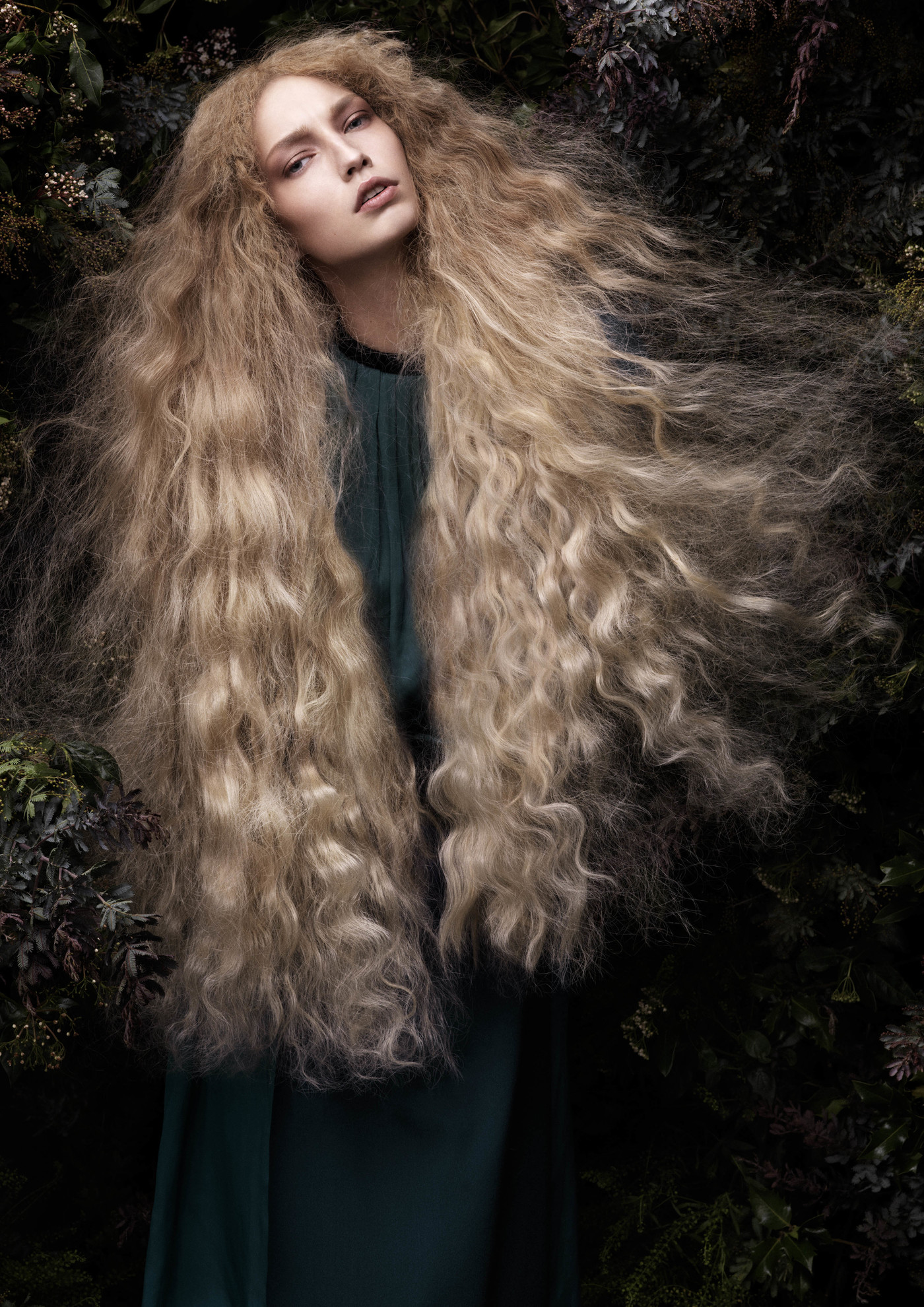Portrait of a fair-skinned woman with voluminous blonde wavy hair, wearing a dark green dress, posing against a dark green leafy background.