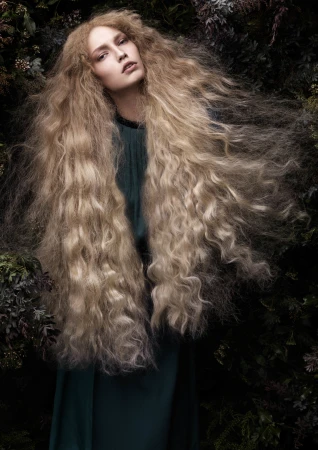 Portrait of a fair-skinned woman with voluminous blonde wavy hair, wearing a dark green dress, posing against a dark green leafy background.