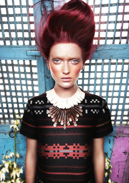 Fashionable portrait of a woman with red hair, wearing a patterned dress and statement jewelry, set against a blue lattice backdrop.