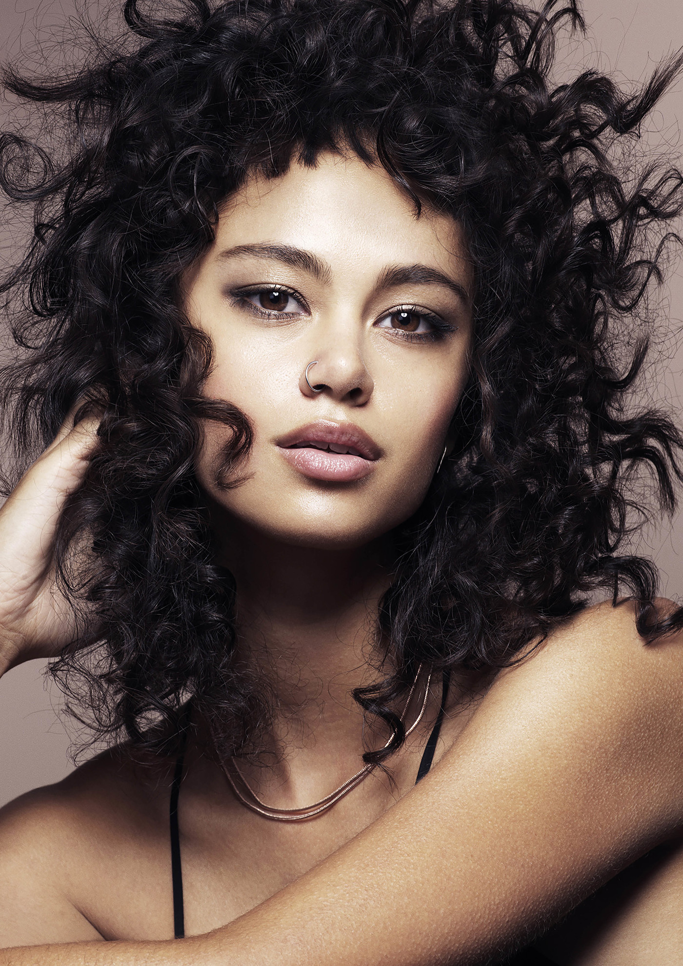 Portrait of a woman with curly hair looking at the camera in a studio. Her bare shoulders are visible. She is wearing a necklace.