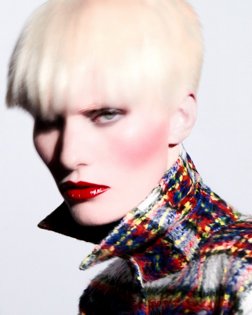 Close-up portrait of a fair-skinned adult wearing a patterned tweed jacket and bold red lipstick against a stark white backdrop.