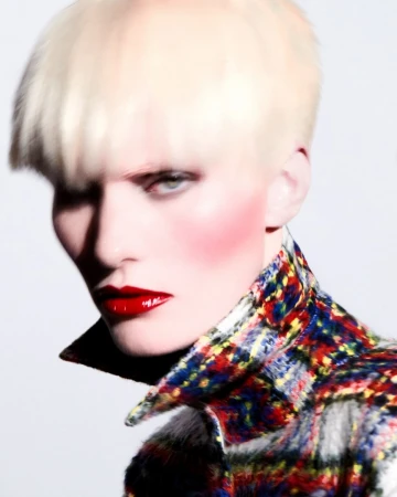 Close-up portrait of a fair-skinned adult wearing a patterned tweed jacket and bold red lipstick against a stark white backdrop.