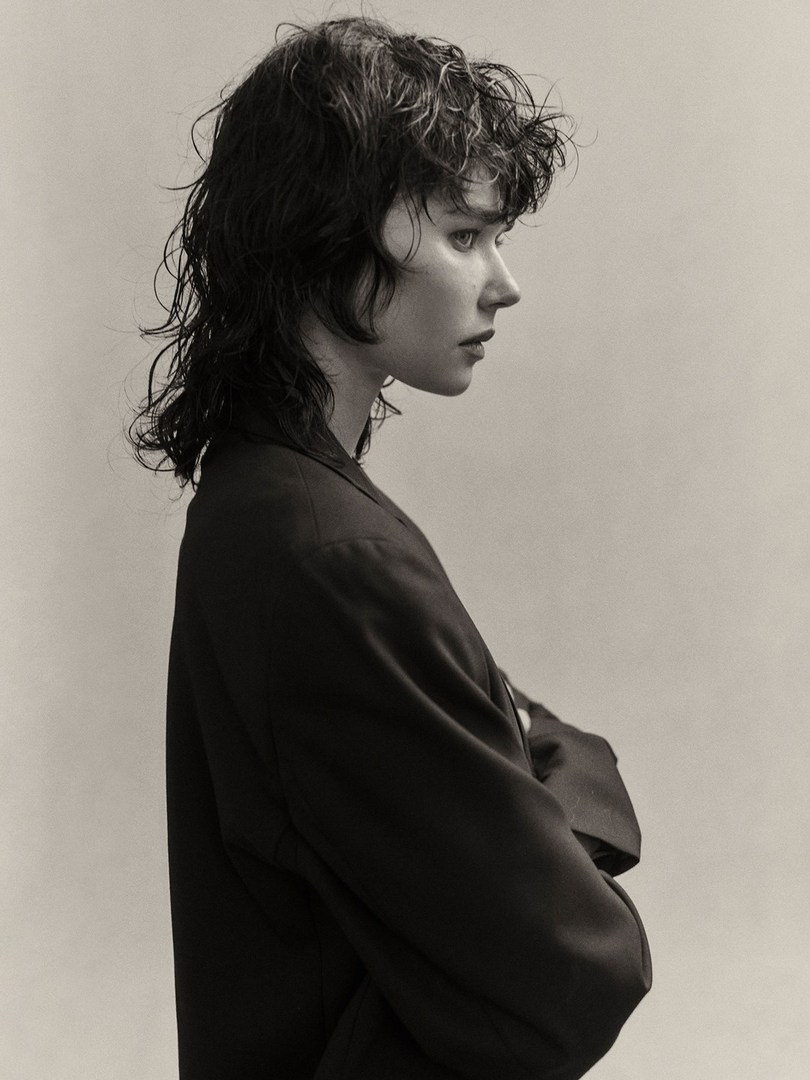 Studio headshot of a young woman with wet wavy hair and dark blazer in profile against a neutral background, showing fashion-forward style.