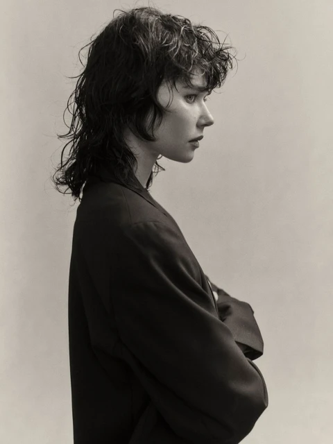 Studio headshot of a young woman with wet wavy hair and dark blazer in profile against a neutral background, showing fashion-forward style.