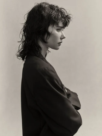 Studio headshot of a young woman with wet wavy hair and dark blazer in profile against a neutral background, showing fashion-forward style.