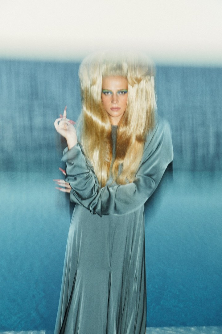 Woman in long turquoise gown with voluminous blonde hair poses against a blurred background in a fashion or beauty shot.
