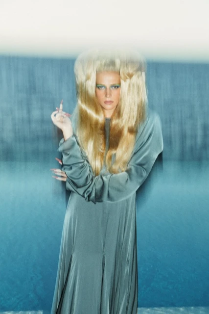 Woman in long turquoise gown with voluminous blonde hair poses against a blurred background in a fashion or beauty shot.