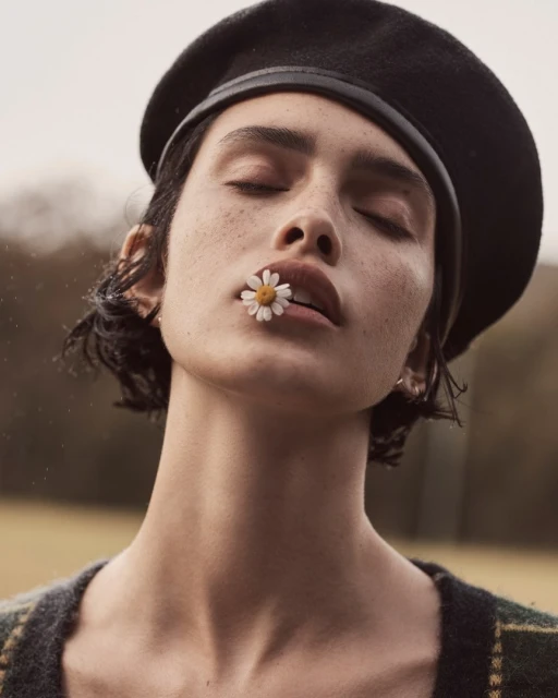 Close-up portrait of a woman wearing a black beret with a daisy in her mouth, eyes closed, in a soft outdoor setting.