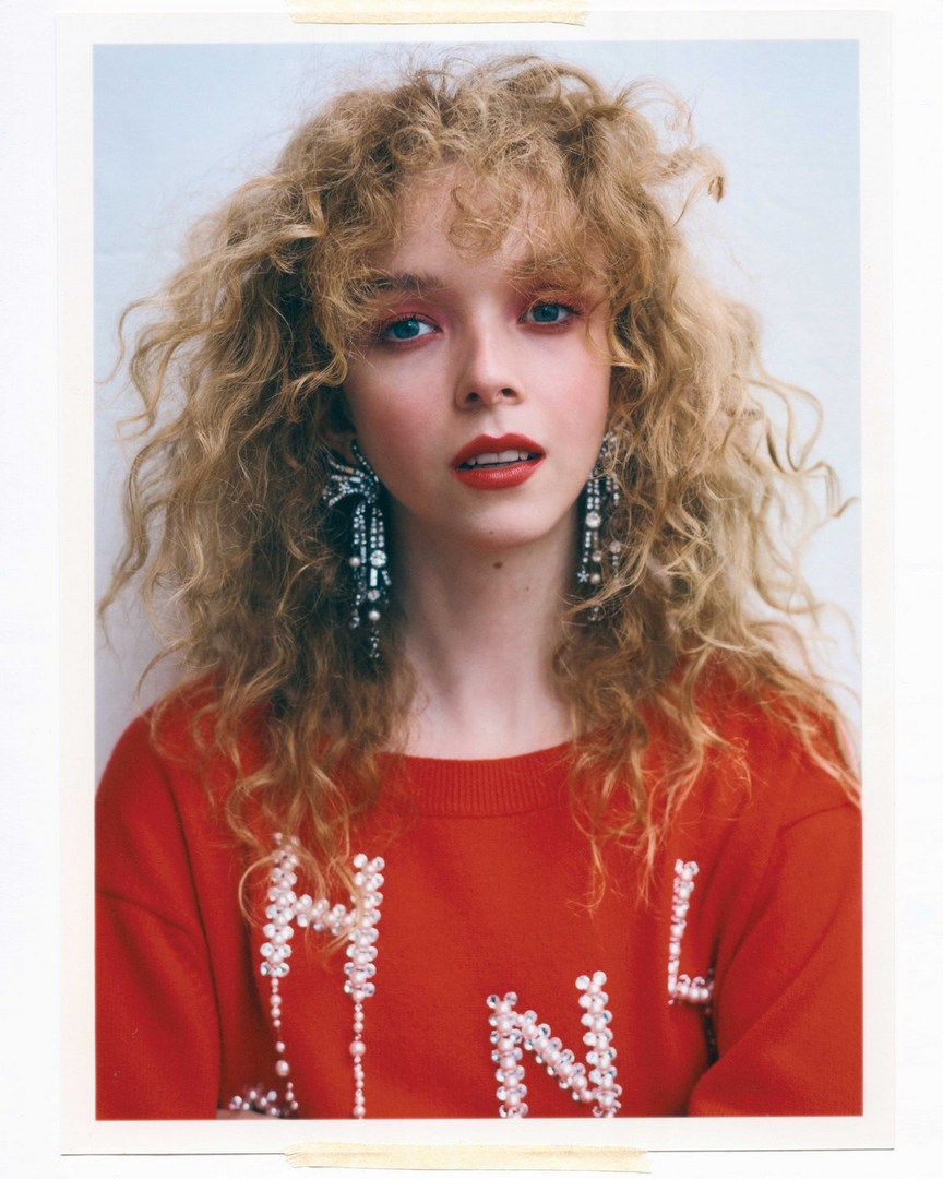 Portrait of a young woman with curly blonde hair, wearing a red sweatshirt and long, dangling earrings, set against a plain white background.