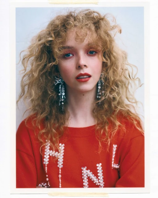 Portrait of a young woman with curly blonde hair, wearing a red sweatshirt and long, dangling earrings, set against a plain white background.