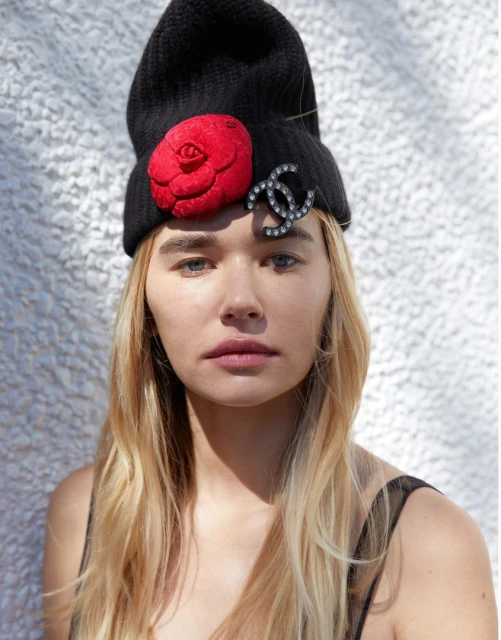 Woman wearing a black beanie hat adorned with a red flower and logo, captured in a medium portrait shot against a neutral background.