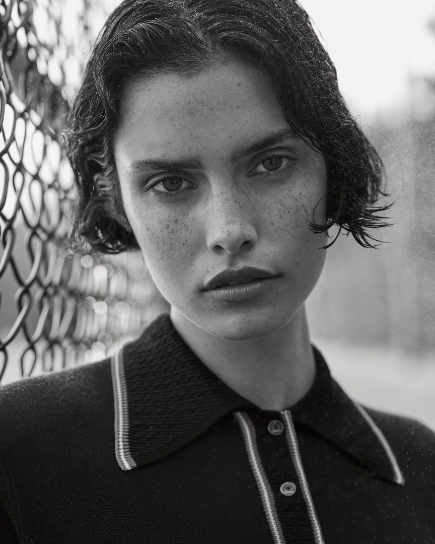 Monochrome portrait of a brunette woman with freckles, looking directly at the camera, wearing a collared shirt, next to a chain-link fence.