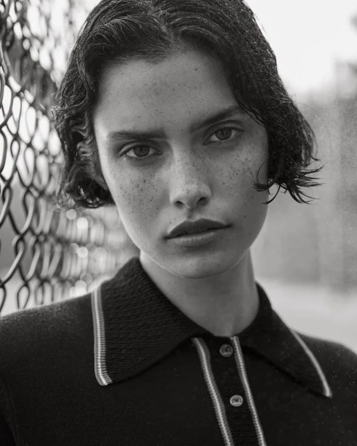 Monochrome portrait of a brunette woman with freckles, looking directly at the camera, wearing a collared shirt, next to a chain-link fence.