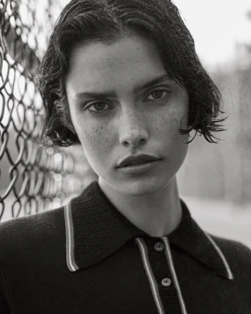 Monochrome portrait of a brunette woman with freckles, looking directly at the camera, wearing a collared shirt, next to a chain-link fence.