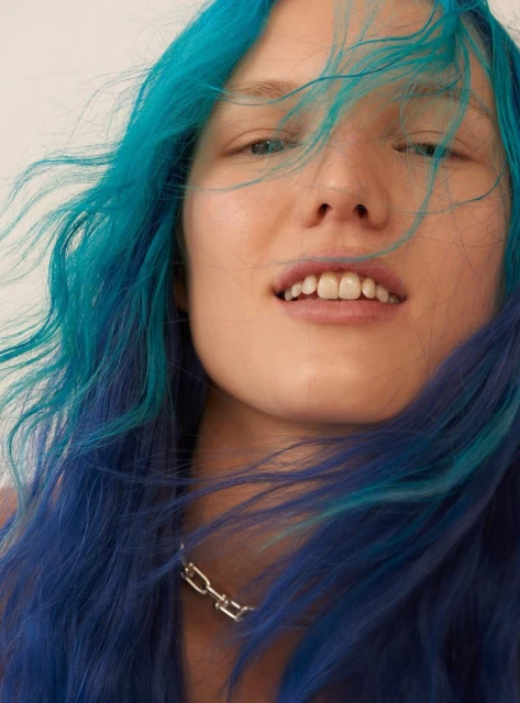 Close-up portrait of a young woman with blue and teal dyed hair and a silver chain necklace against a plain white backdrop.