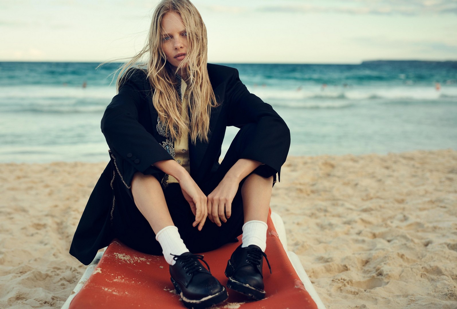 Blonde woman wearing a black jacket and white socks sitting on a red lounger on a sandy beach with the ocean behind her