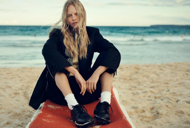 Blonde woman wearing a black jacket and white socks sitting on a red lounger on a sandy beach with the ocean behind her