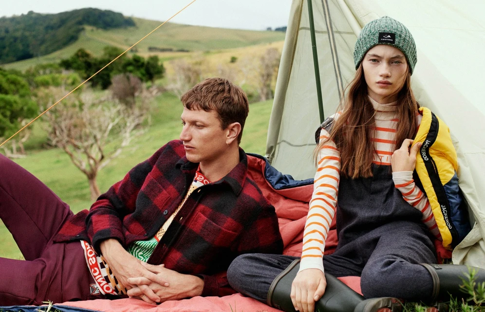 Two people at a campsite: A man in a red and black flannel lies beside a woman in a gray beanie and overalls near a tent.