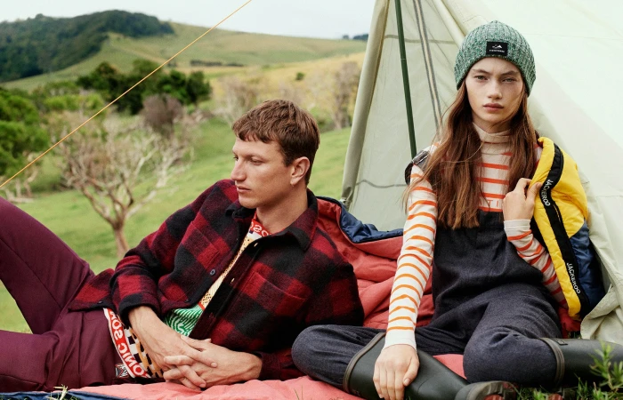 Two people at a campsite: A man in a red and black flannel lies beside a woman in a gray beanie and overalls near a tent.