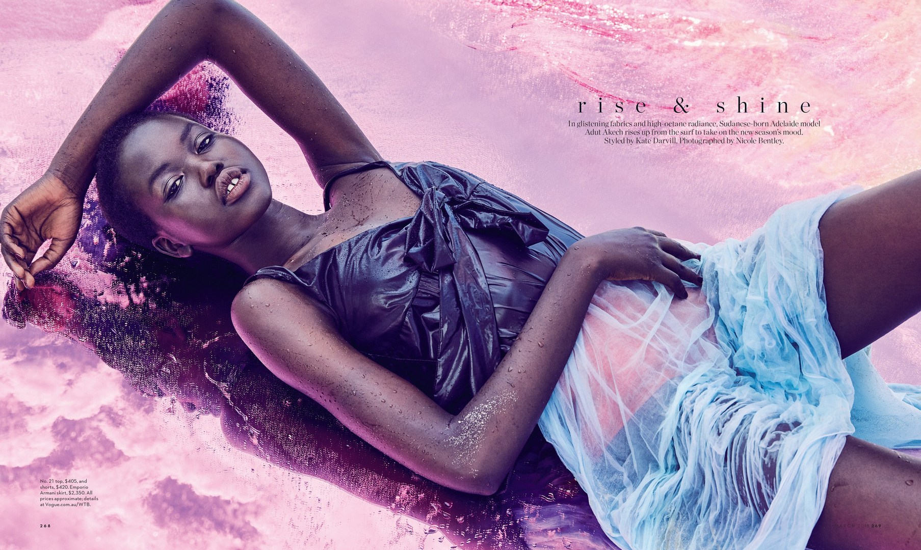 Editorial fashion shot of a dark-skinned woman in a wet black dress and a light blue skirt, lying in pink water, photographed in studio.