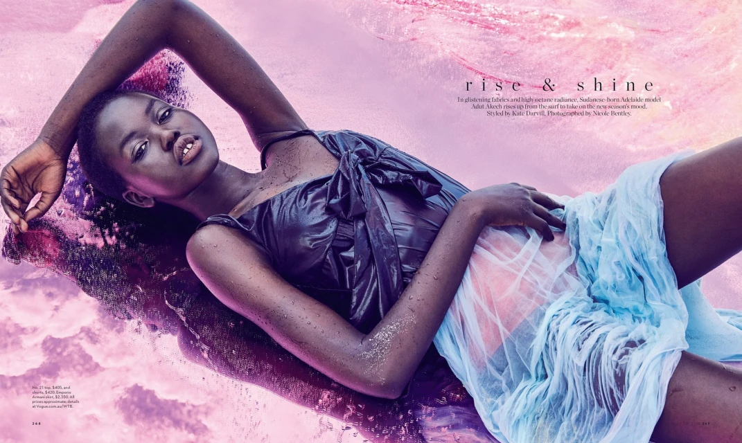 Editorial fashion shot of a dark-skinned woman in a wet black dress and a light blue skirt, lying in pink water, photographed in studio.