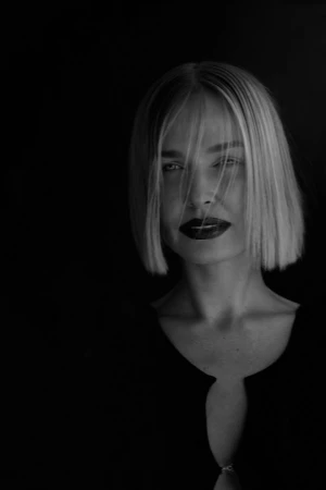Monochrome portrait of a woman with a blonde bob and dark lips wearing a black top against a dark background.
