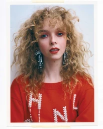 Portrait of a woman with curly blonde hair wearing a red sweater and statement earrings against a white wall