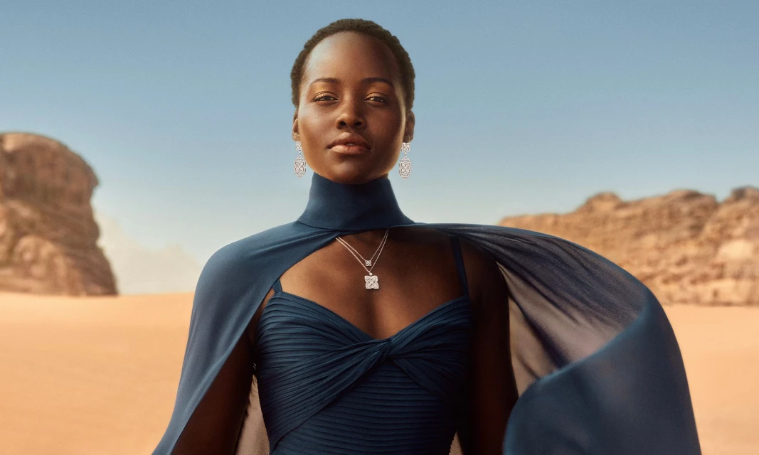 A woman in a blue dress and cape stands in the desert wearing earrings and a necklace, creating a striking and elegant portrait.