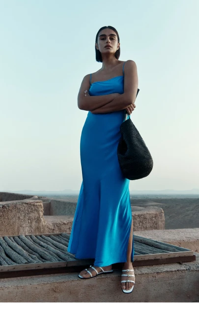 A slender woman in a blue dress with a black bag stands on a rooftop with a tan stone surface and a muted blue sky at dusk.