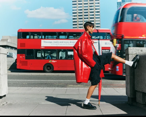 A woman in a red patent leather coat poses on a city street with a red double-decker bus and high-rise buildings in the background.