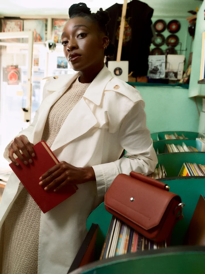 Black woman wearing a white trench coat holding a red book inside a record store with green record bins
