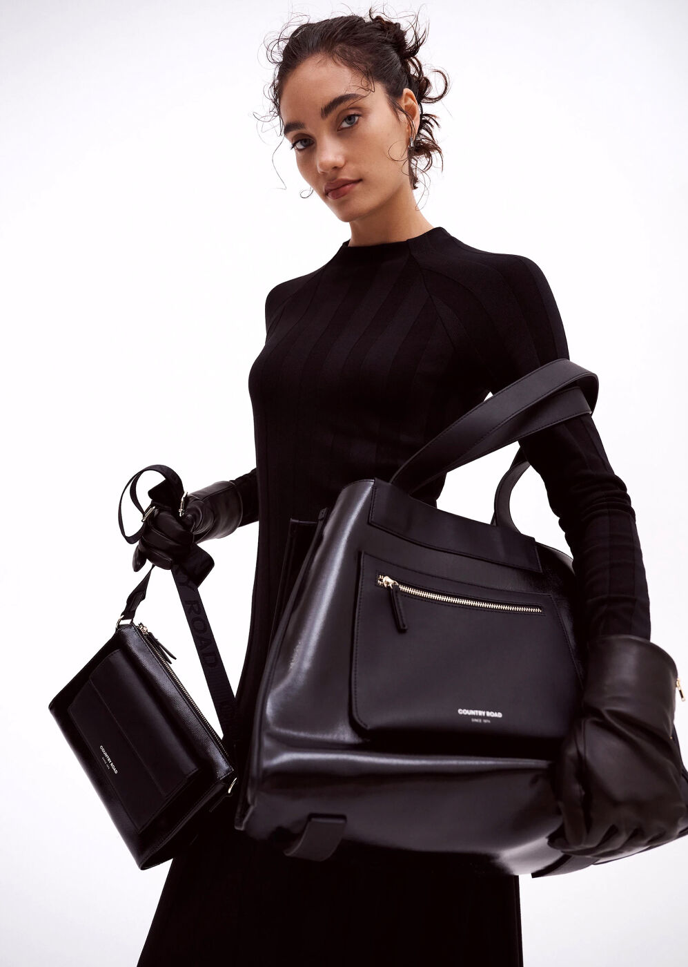 Woman in black dress holding two black leather handbags against a white background in a fashion-forward editorial composition.