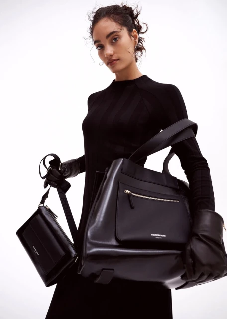 Woman in black dress holding two black leather handbags against a white background in a fashion-forward editorial composition.