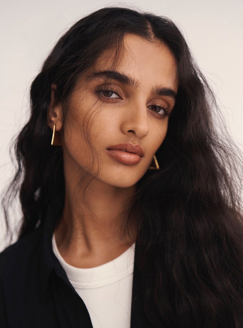Close-up portrait of an adult female model with dark wavy hair wearing a white t-shirt, black collared shirt, and gold earrings.