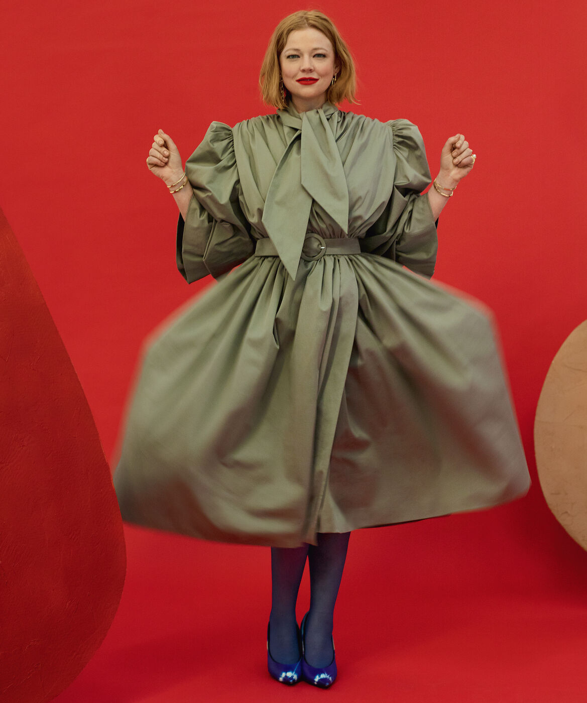 A fair-skinned adult woman with red hair poses in a theatrical, light-green belted dress against a vibrant red backdrop with blue shoes.