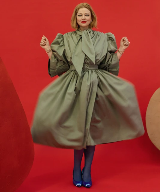 A fair-skinned adult woman with red hair poses in a theatrical, light-green belted dress against a vibrant red backdrop with blue shoes.
