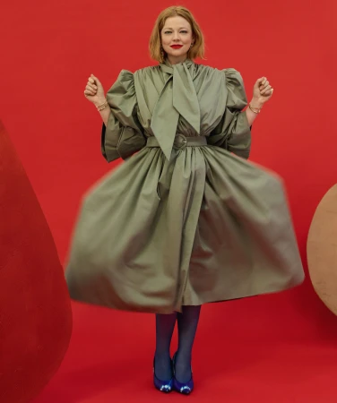 A fair-skinned adult woman with red hair poses in a theatrical, light-green belted dress against a vibrant red backdrop with blue shoes.