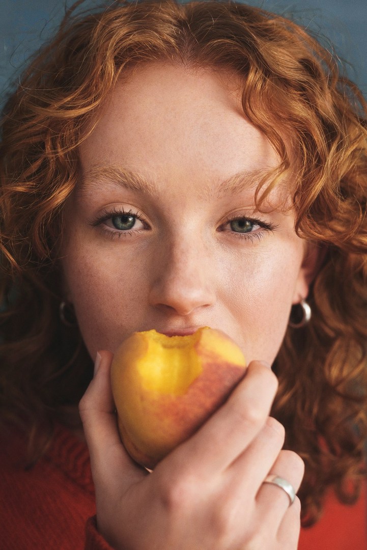 Close-up portrait of a woman with curly red hair holding a fresh bitten peach near her face by Lok Lau