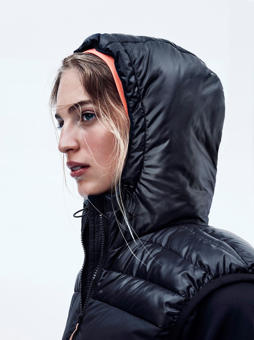 Close-up of a blonde woman with blue eyes wearing a black puffer vest and coral headband against a white background in a studio.