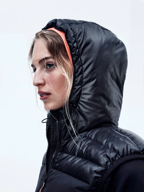 Close-up of a blonde woman with blue eyes wearing a black puffer vest and coral headband against a white background in a studio.