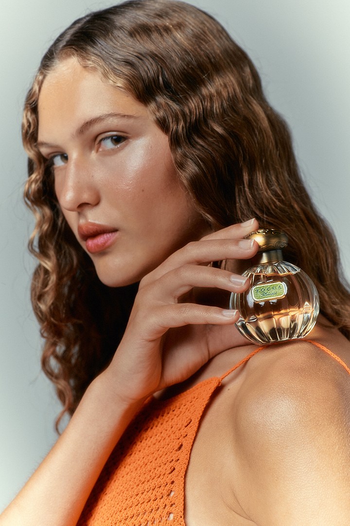 Elegant portrait of an adult woman holding a perfume bottle. She wears an orange top and has wavy brown hair, with a clean, neutral background.