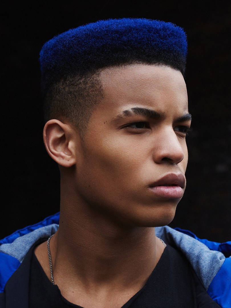 Close-up of a young adult male with a high-top fade haircut dyed vibrant blue, wearing a blue and black jacket and silver chain necklace.