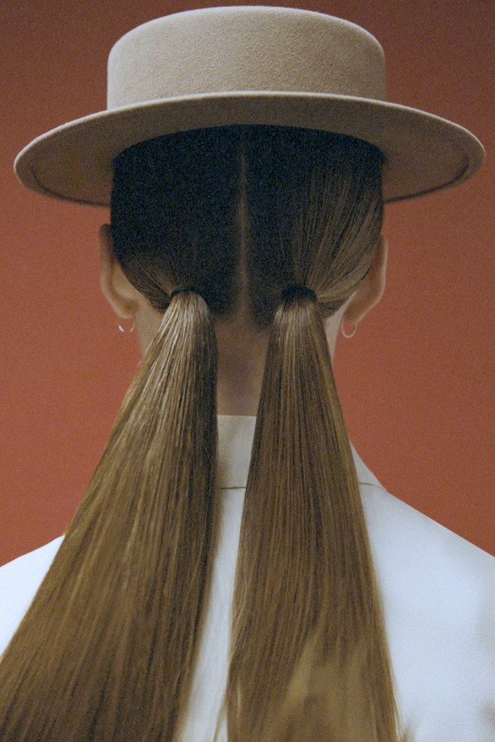 Back view of a woman with long blonde hair in pigtails wearing a tan hat and white top against a red-orange backdrop.