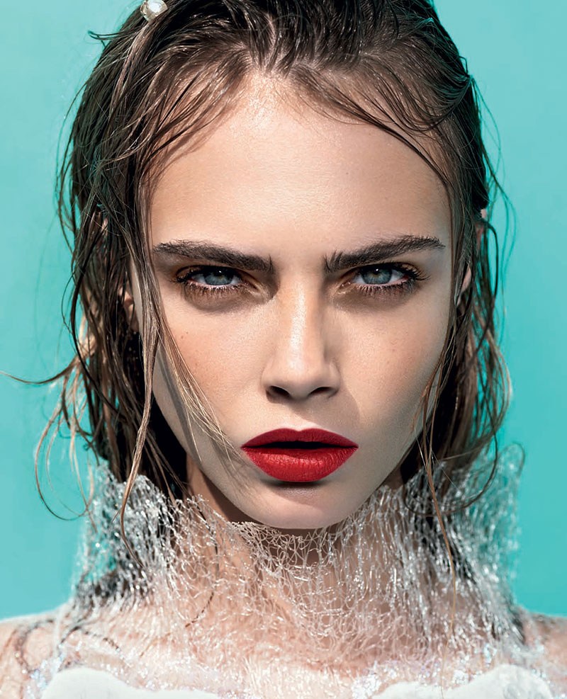 Close-up studio shot of a fair-skinned woman with red lipstick, wet hair, and striking blue eyes against an aqua backdrop, showcasing beauty and fashion.