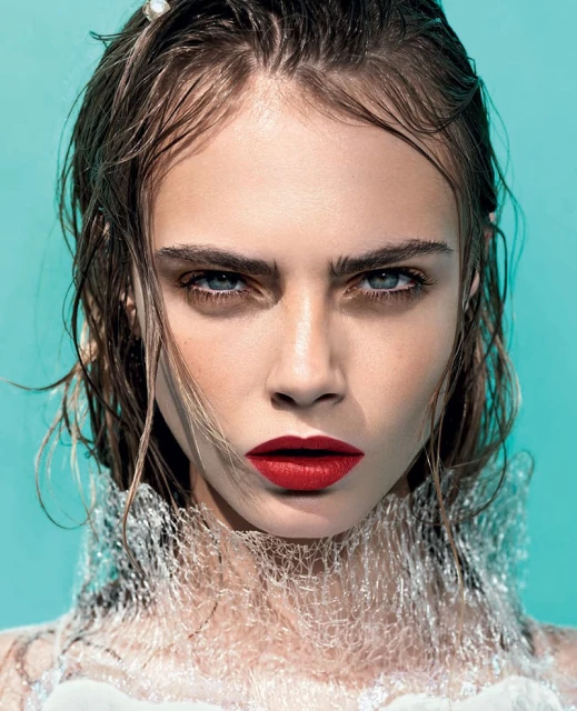 Close-up studio shot of a fair-skinned woman with red lipstick, wet hair, and striking blue eyes against an aqua backdrop, showcasing beauty and fashion.