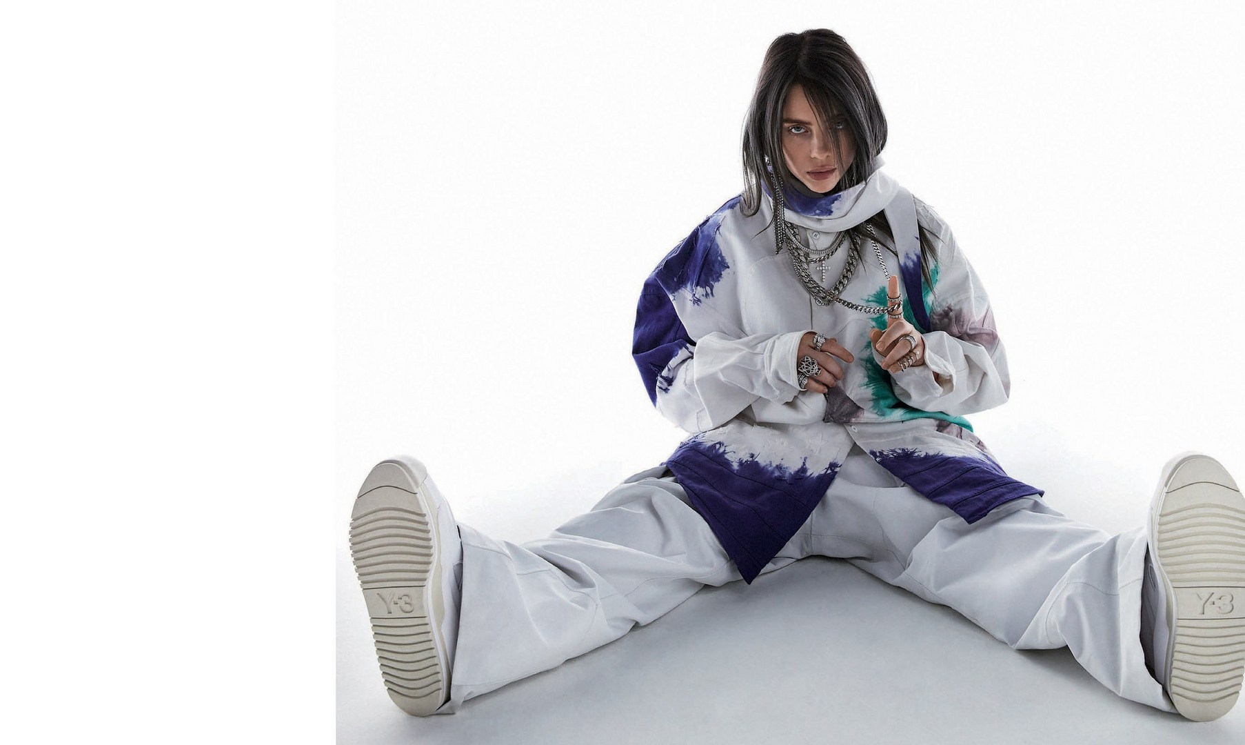 Billie Eilish sitting on a white background wearing an oversized white and purple tie-dye shirt, white trousers, and chunky sneakers.
