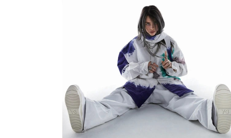 Billie Eilish sitting on a white background wearing an oversized white and purple tie-dye shirt, white trousers, and chunky sneakers.