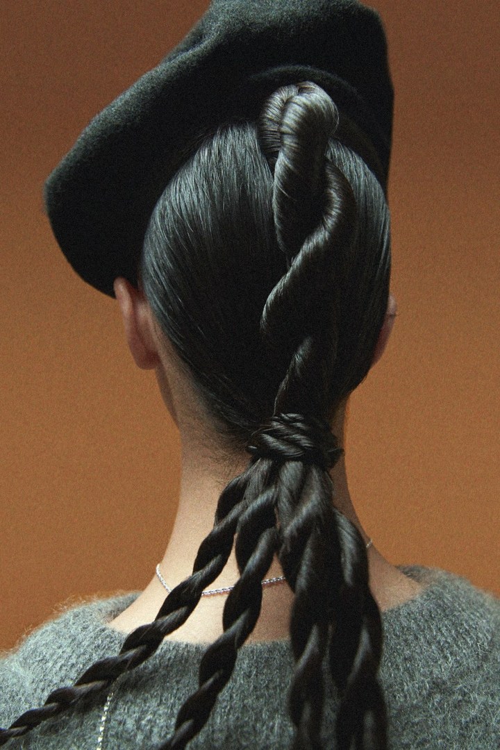 Back view of a person with an elaborate braided hairstyle, wearing a gray sweater and black beret against a warm orange background.
