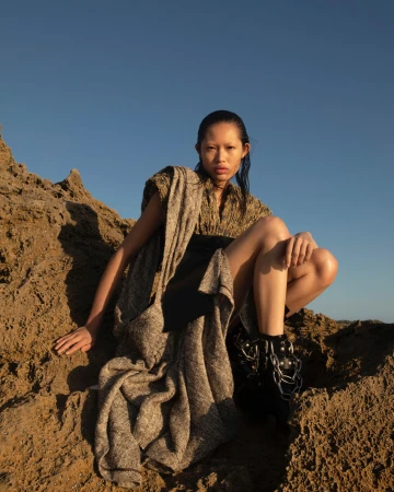 Model crouched on rocky terrain wearing a textured textured garment against a clear blue sky by Lok Lau