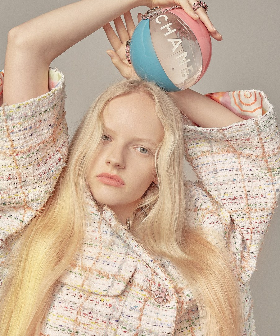 Woman with blond hair holds a Chanel-branded, two-tone pink-and-blue, spherical handbag with a chain strap indoors against a neutral background.